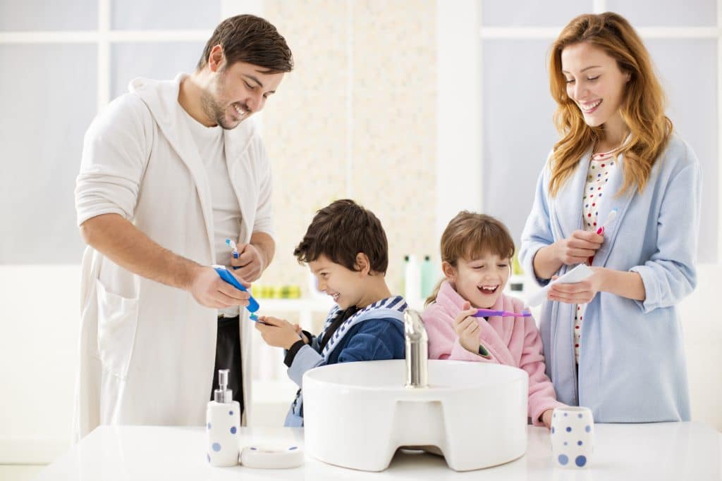 Portrait of a happy family with two children brushing teeth together at home in a bathroom. Father and mother applying toothpaste to children's toothbrush.